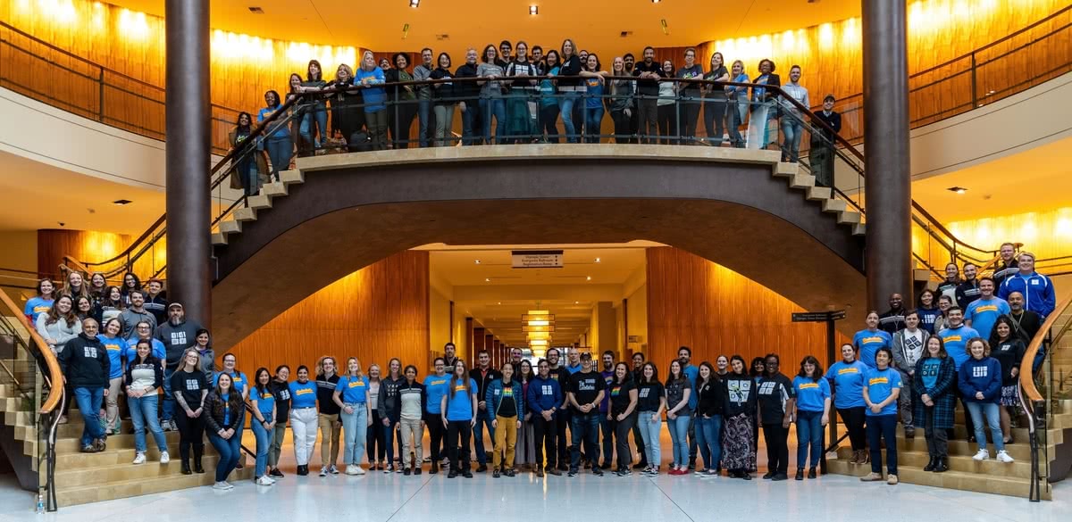 Nearly 100 Code.org employees in matching blue and yellow t-shirts pose in a hotel conference room for the company's summer 2022 Codechella event.